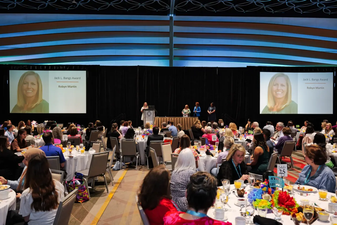 Audience seated at an awards breakfast beneath a stage screen.