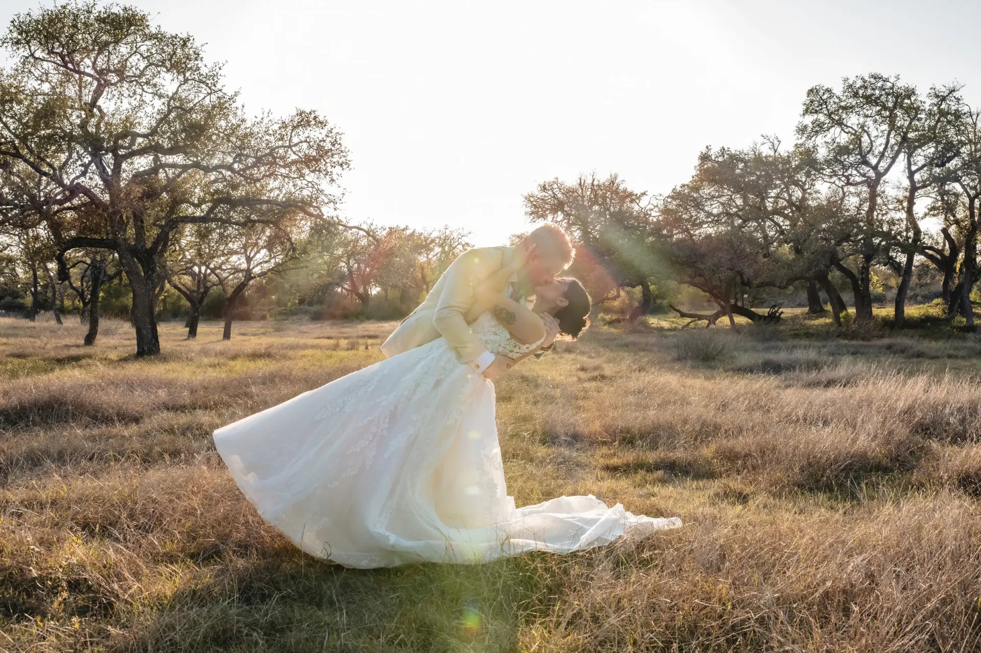 Newlyweds share a dip beneath oak trees in warm backlight.