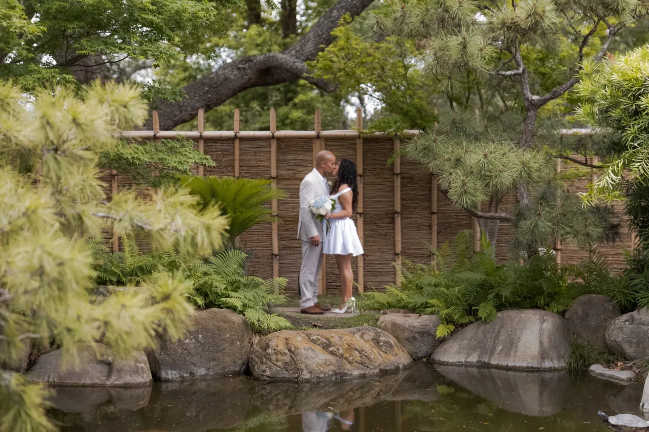 Newlyweds stand together beside a garden pond.