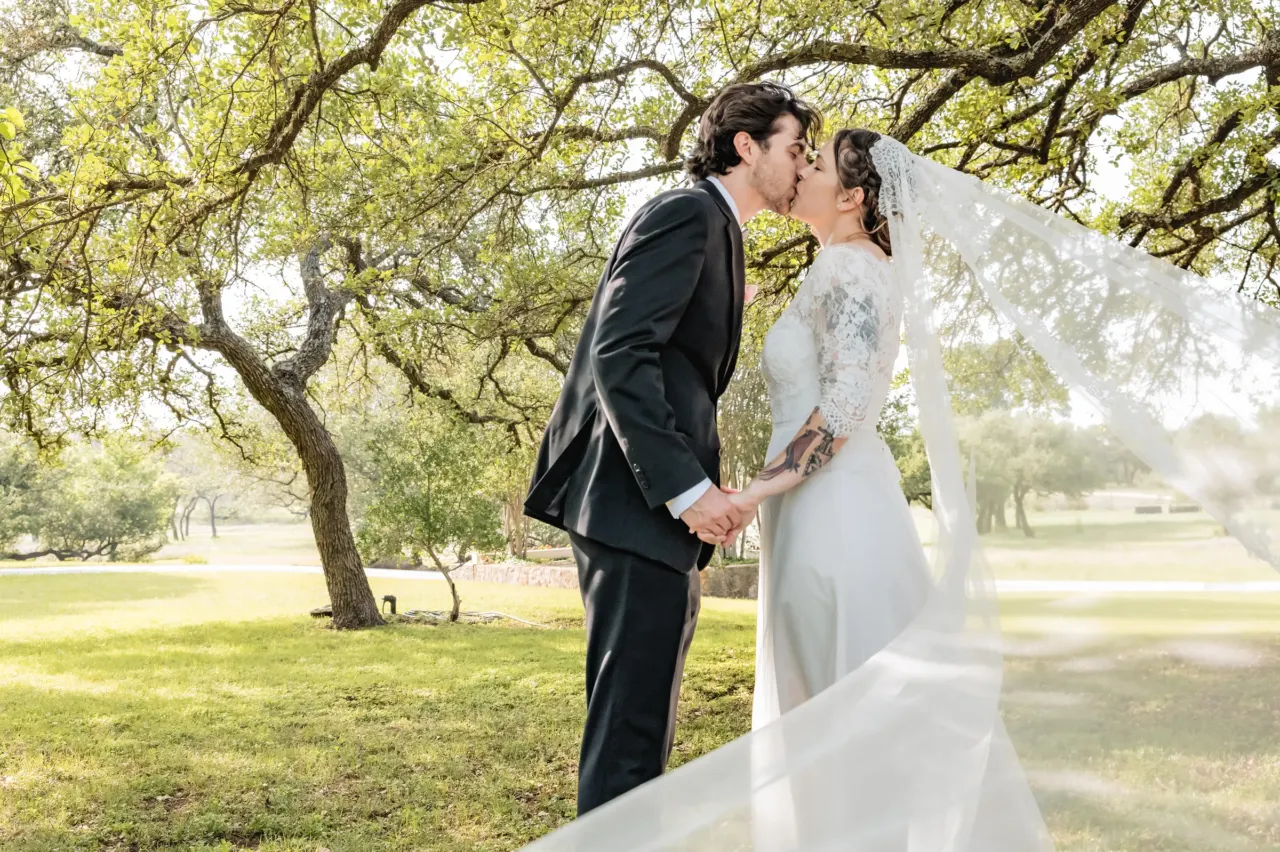 Newlyweds kiss beneath oak trees in a sunlit park.