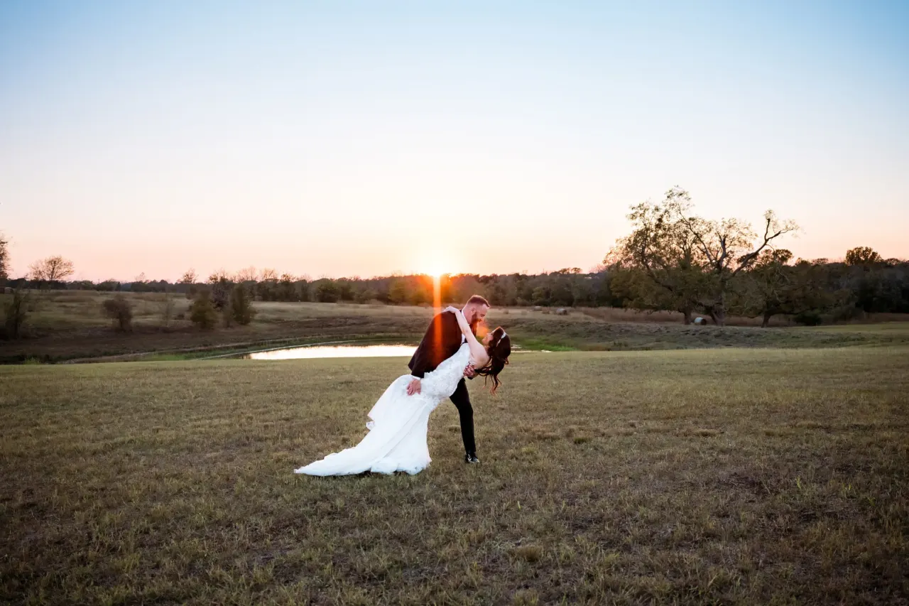 Newlyweds share a dip and kiss in a sunset field.